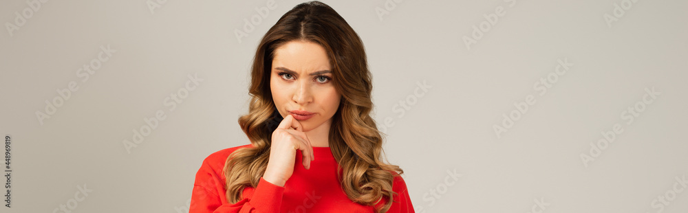 thoughtful curly woman in red sweatshirt looking at camera isolated on grey, banner thoughtful curly woman in red sweatshirt looking at camera isolated on grey, banner