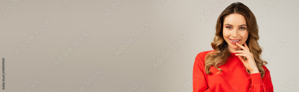 happy woman in sweatshirt looking at camera while touching lips isolated on grey, banner happy woman in sweatshirt looking at camera while touching lips isolated on grey, banner