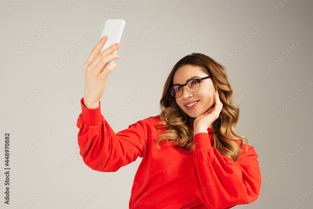 joyful woman in eyeglasses taking selfie on smartphone isolated on grey joyful woman in eyeglasses taking selfie on smartphone isolated on grey