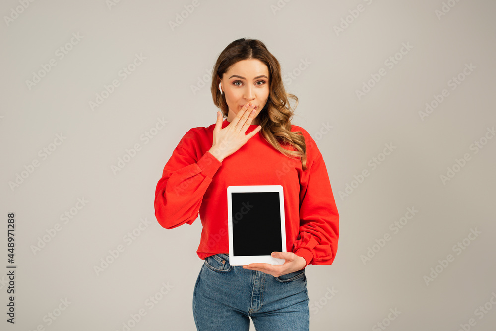 Fototapeta premium surprised woman in wireless earphones holding digital tablet with blank screen isolated on grey