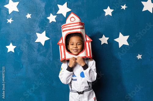 Rocket afro-american little boy in space suit playing astronaut on blue background with white hand-made stars. Happy mixed race child with handcrafted spacecraft. Childhood, creative and imagination.