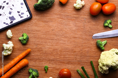 Top view. Cutting boards, knives, and a variety of vegetables. Tomatoes, carrots, scallions, and broccoli form a circle on a wooden table with space in the middle for captions.