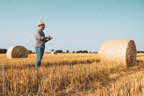 Happy farmer is standing beside bales of hay. He is examining straw after successful harvesting. © djoronimo