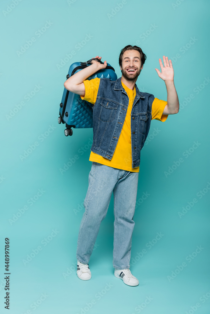 Happy traveler in denim vest holding suitcase and waving hand on blue background Happy traveler in denim vest holding suitcase and waving hand on blue background