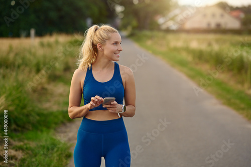 Fit athletic woman pausing to consult her mobile phone while out jogging on a rural footpath looking to the side with a happy smile of satisfaction and pleasure