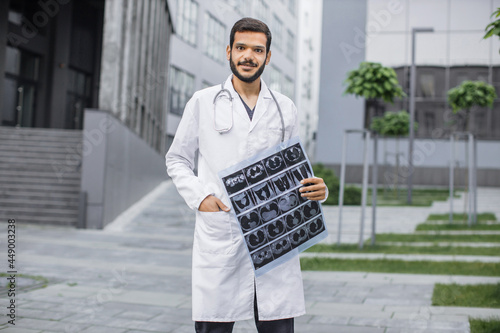 Handsome male Arab doctor with stethoscope, wearing white coat, looking at camera with smile, holding tomography scan, standing outdoors near clinic. Medical and radiology concept © sofiko14