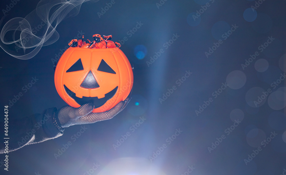 Fototapeta premium A bucket of candy in the form of a pumpkin lantern Jack on a black background in a hand. Halloween banner place for text.
