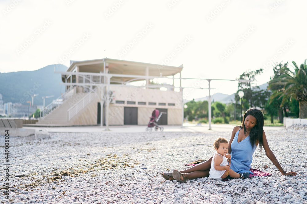Little girl sitting next to her mother on a pebble beach against the backdrop of a building