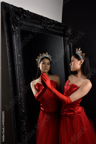 Full length portrait of beautiful young asian woman wearing red corset, long opera gloves and ornate crown headdress. Graceful posing against a full length mirror with a dark studio background.