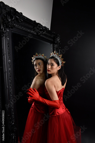 Full length portrait of beautiful young asian woman wearing red corset, long opera gloves and ornate crown headdress. Graceful posing against a full length mirror with a dark studio background.