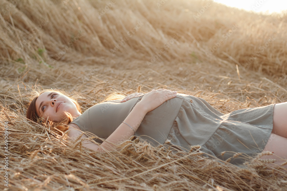 Caucasian pregnant young red-haired woman in dress lying on wheat field in summer at sunset, future mother relaxing in nature