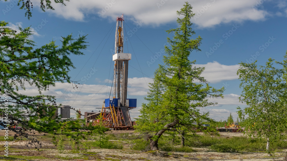 Landscape with a drilling rig in an oil and gas field. Infrastructure, communications and drilling equipment are visible. Around the rig there is summer northern taiga with green trees and blue sky