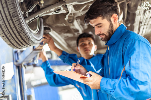 Two professional look technician inspecting car underbody and suspension system by using check list in moder car service shop. Automotive business or car repair concept.	