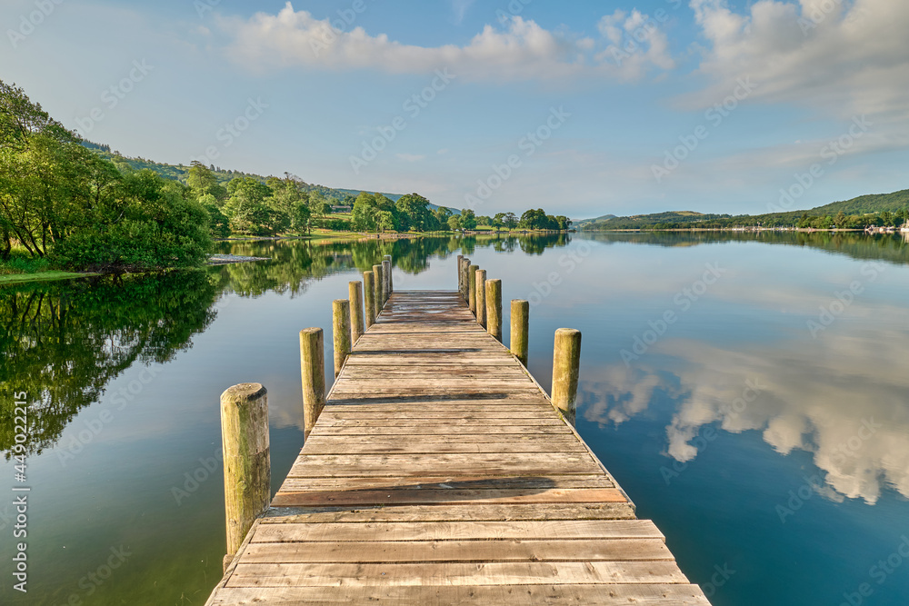 A wooden jetty on Coniston Water in the Lake District, shot in landscape, looking straight down the jetty to the hills beyond, with clouds reflected in the water