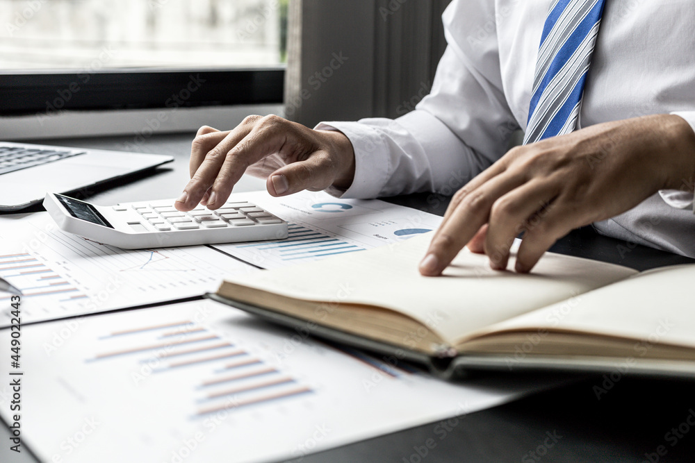 Obraz premium Businessman is pressing a calculator and looking at the information in his notebook, he is checking the balance from the monthly financial and sales documents. Concept of finance and sales management.