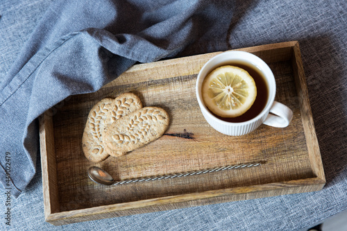 A white cup of tea with lemon, a long cupronickel spoon with a twisted handle and a saucer with three oatmeal cookies for breakfast on a wooden tray, a rag napkin on a gray sofa. photo