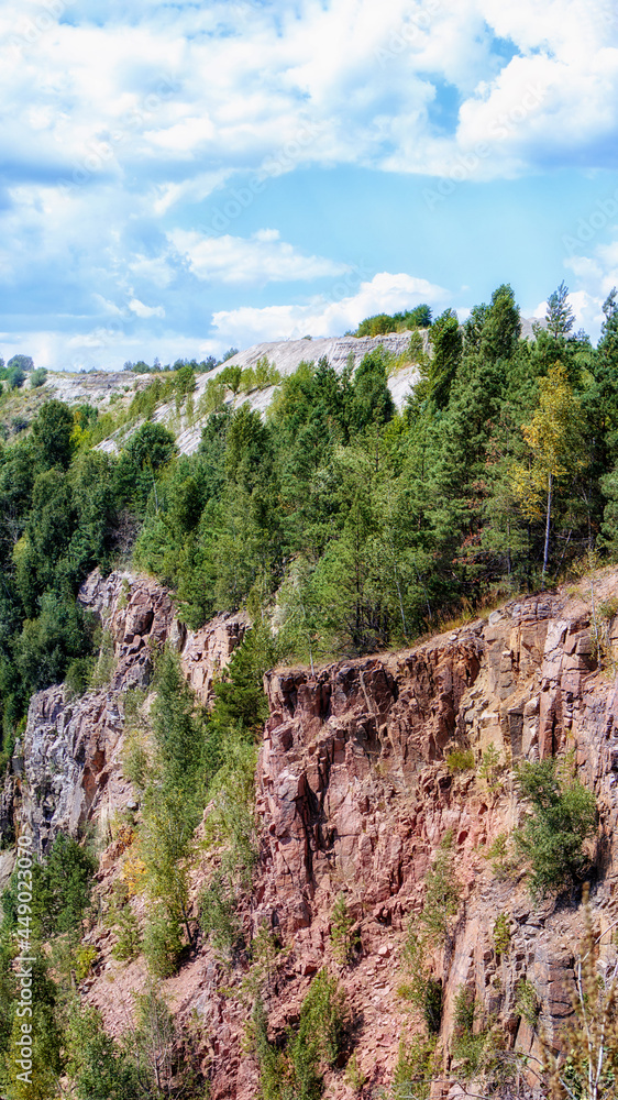 Abandoned granite quarry in Ukraine, vertical panorama. Rocky slope of quarry with sharp stones overgrown with trees. Summer landscape with cliffs and cloudy sky.