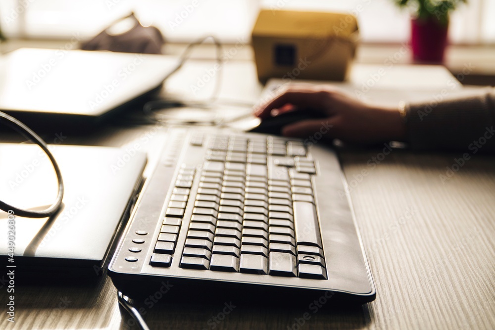 A portrait of a computer keyboard lying on a wooden table with someone using a mouse in the blurry background, the person is using the pc in an indoor environment.