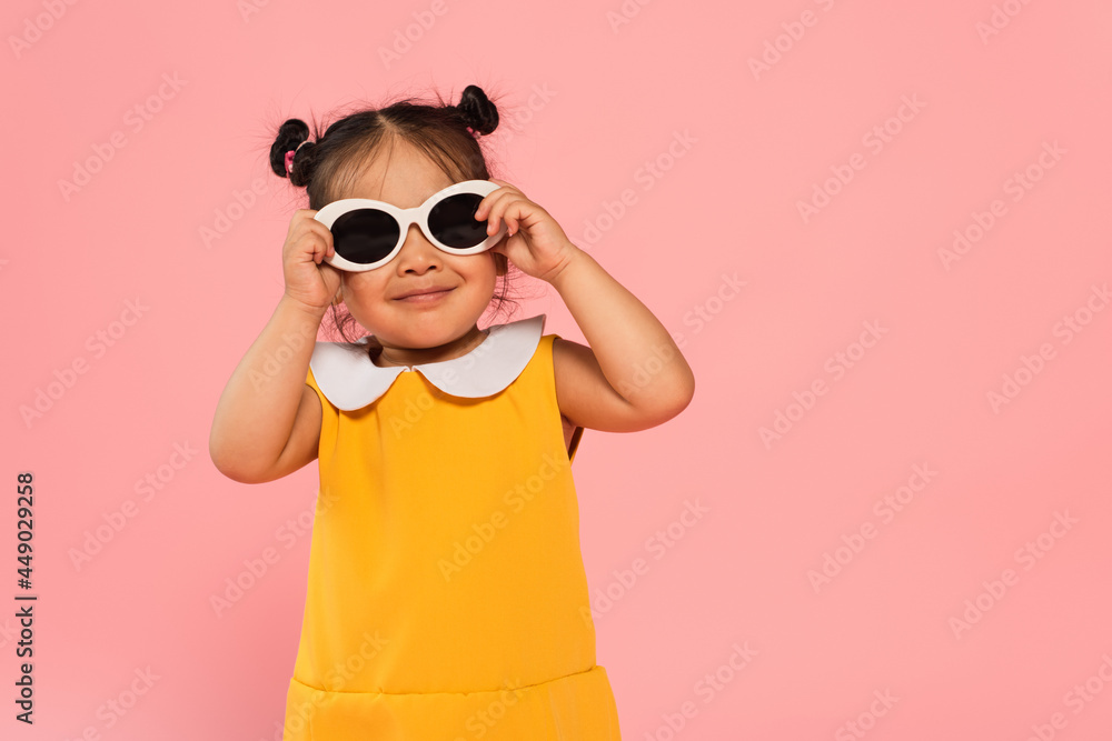 cheerful asian toddler kid in yellow dress adjusting sunglasses isolated on pink cheerful asian toddler kid in yellow dress adjusting sunglasses isolated on pink