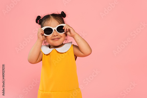 cheerful asian toddler kid in yellow dress adjusting sunglasses isolated on pink