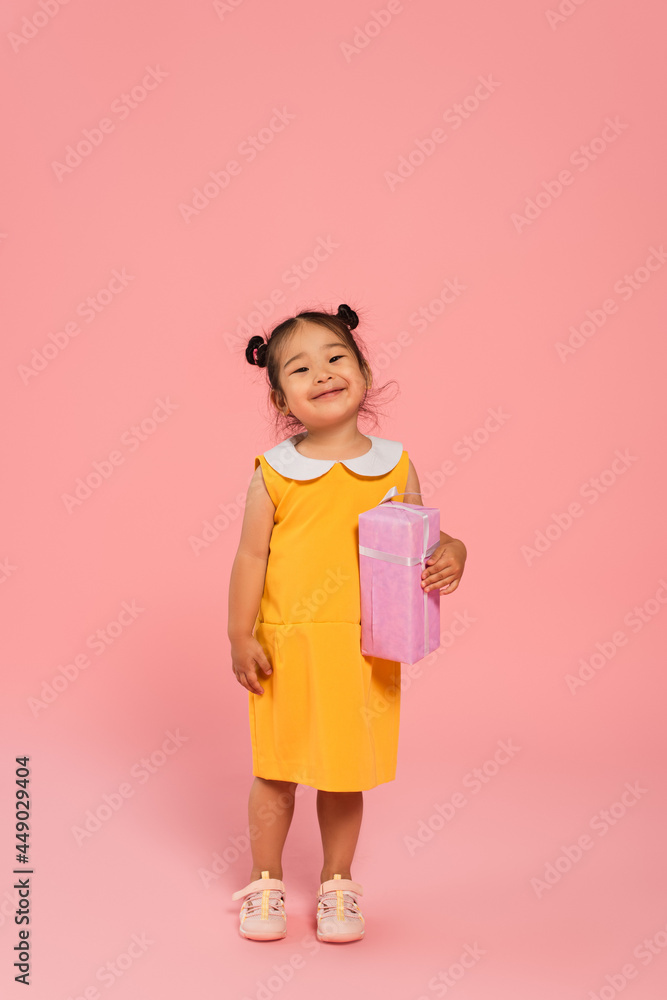 full length of pleased asian toddler kid in yellow dress holding present on pink full length of pleased asian toddler kid in yellow dress holding present on pink