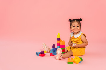 happy asian toddler girl in yellow dress playing building blocks on pink