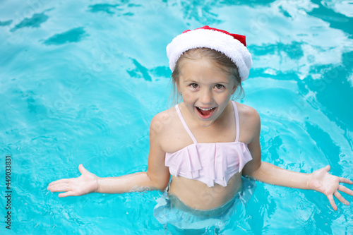 a beautiful happy girl in a Santa Claus hat in a pink swimsuit is standing in the pool for the new year