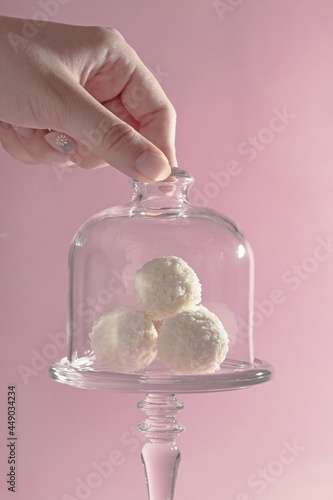 round sweet candies in coconut shavings in a glass vase on a pink background, harmony of taste, exquisite dessert, hand takes rafaello photo