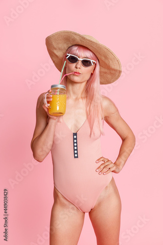 Attractive fashionable woman wearing straw hat, swimsuit and sunglasses drinking tropical cocktail, posing isolated over pink studio background. Summer vacation concept