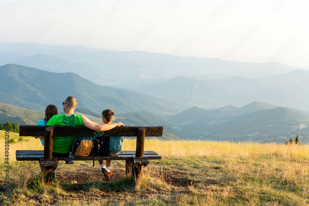 Cheerful father playing with his children's on the bench in meadow at mountain. Father and children sitting on the bench having relaxing conversation. Cheerful father playing with his children's on the bench in meadow at mountain. Father and children sitting on the bench having relaxing conversation.