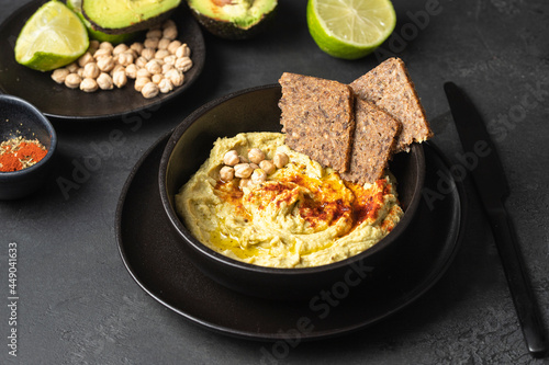 Black ceramic bowl with chickpea hummus on black background with chickpea beans , avocado, smoked paprika and multigrain bread
