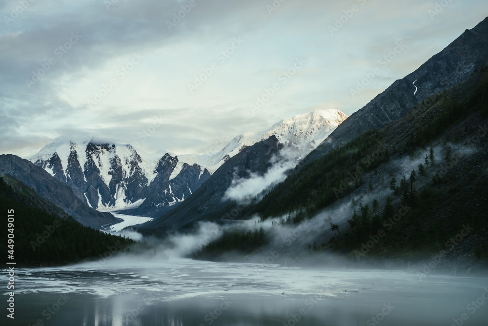Scenic alpine landscape with snowy mountain peak in golden sunlight and mountain lake in fog under cloudy sky. Atmospheric highland scenery with sunlit mountain top and low clouds on rocks and slopes.