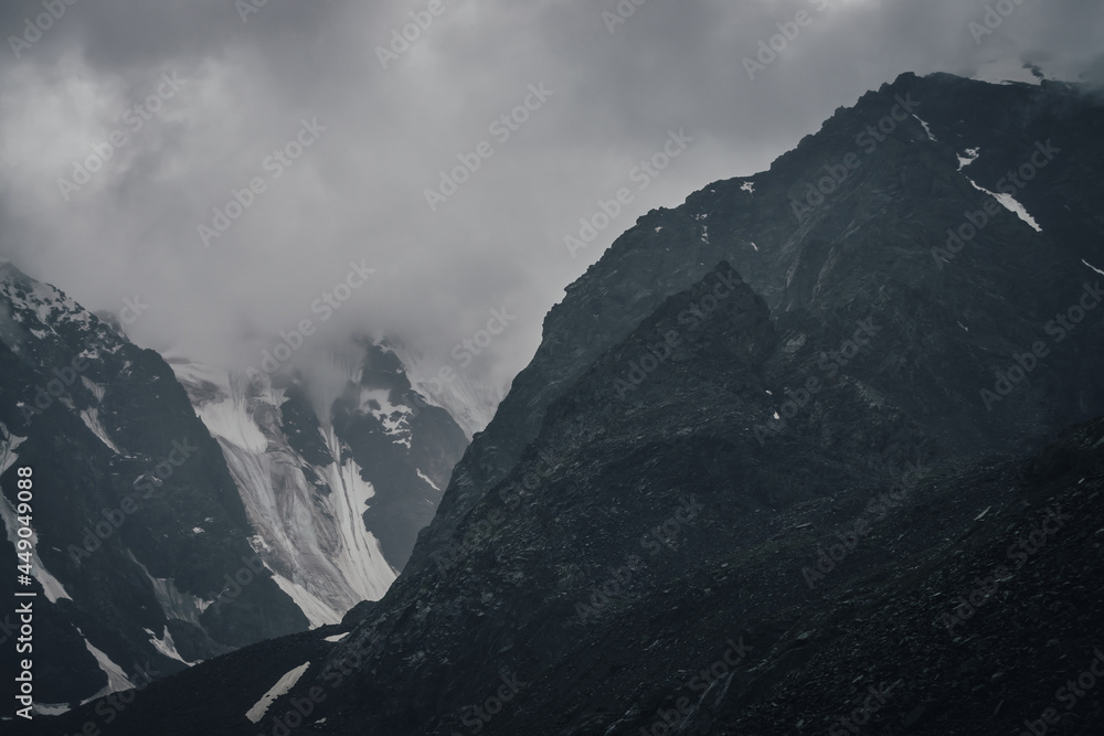 Dark atmospheric mountain landscape with glacier on black rocks in lead gray cloudy sky. Snowy mountains in low clouds in rainy weather. Gloomy landscape with black rocky mountains with snow in fog.