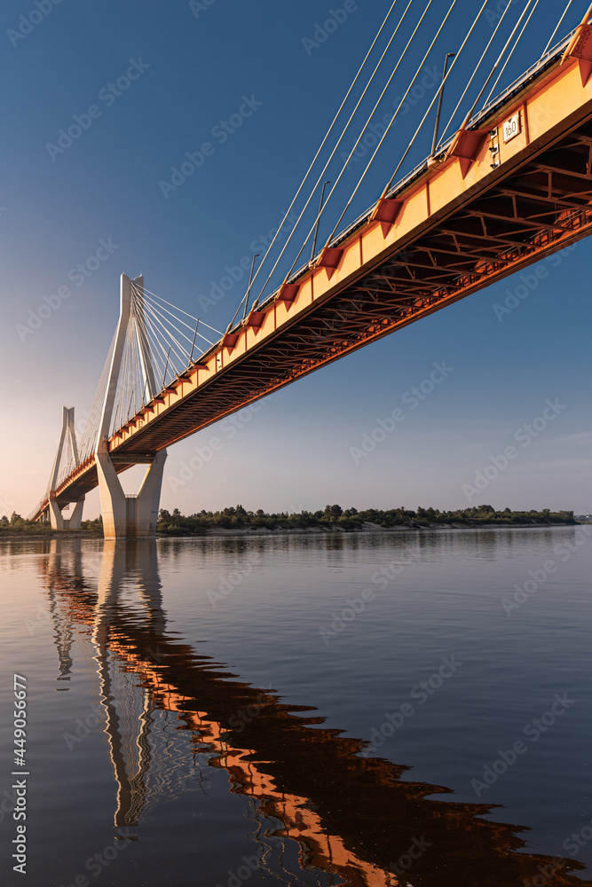 Fototapeta premium A view from the ground to a suspension bridge with two huge pillars reflecting in a river at sunrise in a rural area