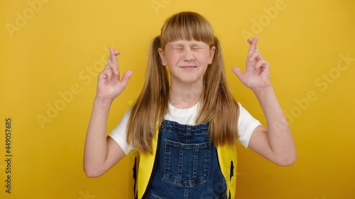 Portrait cute blonde kid schoolgirl keep fingers crossed, wears backpack posing isolated over yellow color background wall in studio. People childhood lifestyle concept wait for special moment 