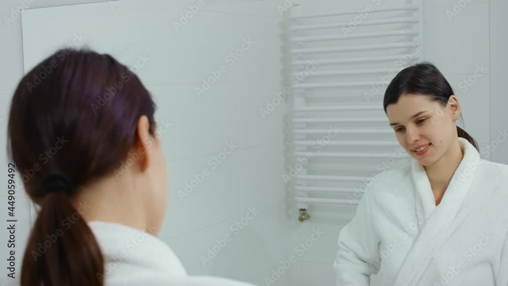 Front view of young woman coming out of shower and standing in bathrobe in front mirror admiring herself in bathroom. Concept of woman admiring herself in mirror. Front view of young woman coming out of shower and standing in bathrobe in front mirror admiring herself in bathroom. Concept of woman admiring herself in mirror.