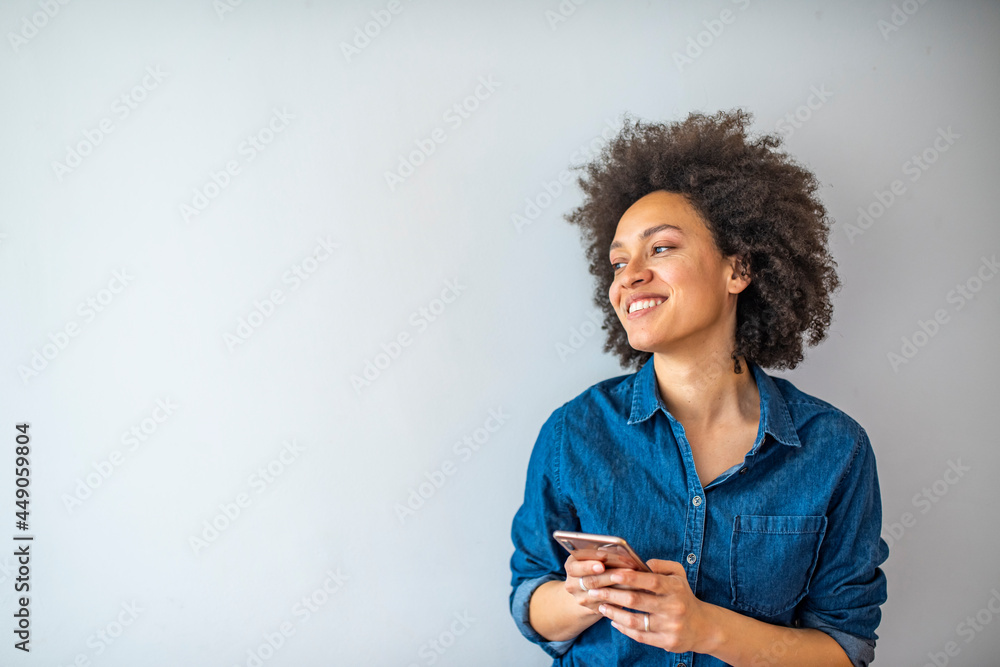 Smiling mature woman using smartphone isolated on gray wall with copy space. Happy African American woman in casual typing on cellphone over grey background. Smiling mature woman using smartphone isolated on gray wall with copy space. Happy African American woman in casual typing on cellphone over grey background.