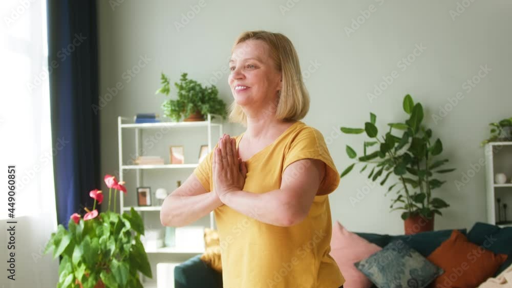 Elderly smiling woman standing on gym mat and doing yoga exercises in living room. Female pensioner practicing pilates on floor, relaxing and meditating in morning. Remote sports training Elderly smiling woman standing on gym mat and doing yoga exercises in living room. Female pensioner practicing pilates on floor, relaxing and meditating in morning. Remote sports training