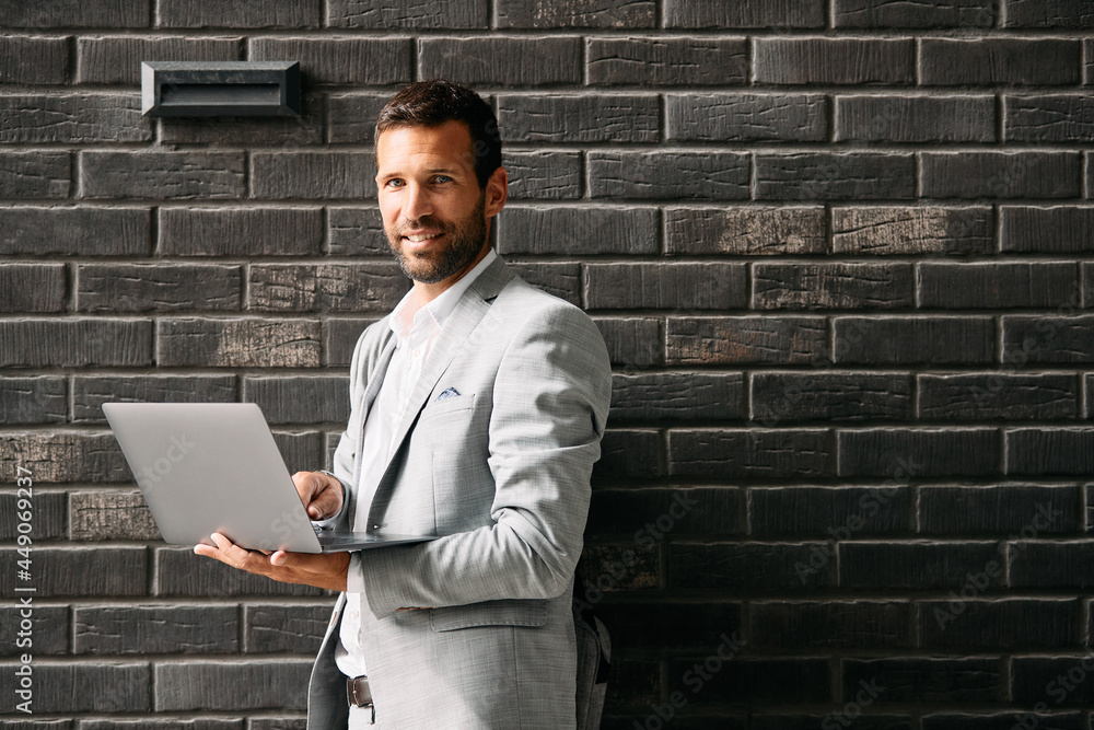Smiling businessman uses laptop against the wall while looking at camera.