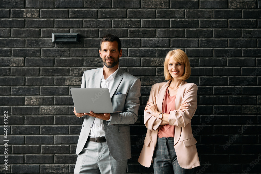 Happy business colleagues with laptop standing against the wall and looking at camera. Happy business colleagues with laptop standing against the wall and looking at camera.