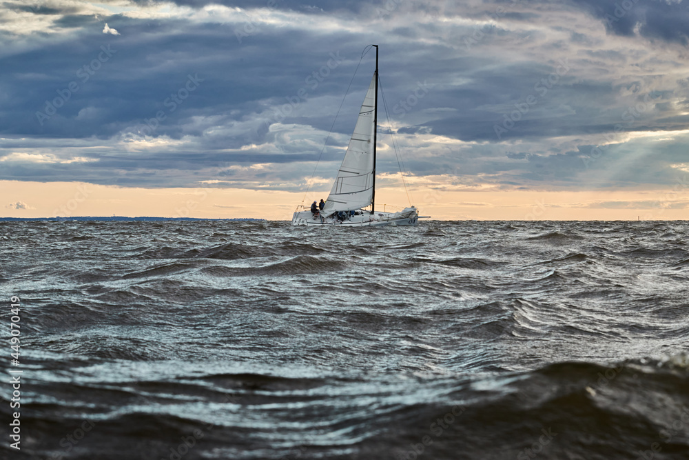 The lonely sailboat on the horizon in sea at sunset, the storm sky of different colors, big waves, sail regatta, cloudy weather, only main sail, sun beams