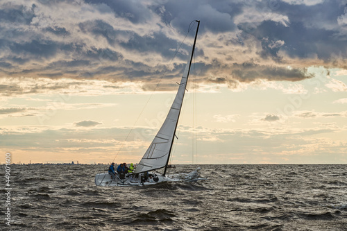 Russia, St. Petersburg, 23 July 2021: The lonely sailboat on the horizon in sea at sunset, the storm sky of different colors, big waves, sail regatta, cloudy weather, only main sail, sun beams