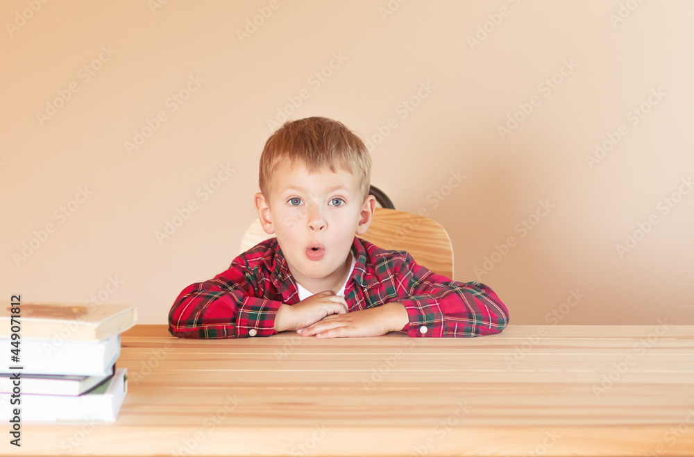Cute little boy sits at a desk. He is surprised and widens his eyes. Back to school concept Cute little boy sits at a desk. He is surprised and widens his eyes. Back to school concept