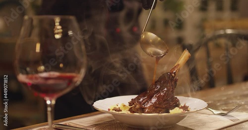 Spoon pouring sauce on meat. Cooked lamb knuckle . Dark red sauce poured on meat of lamb in plate with salad . Chef's Table with glass of wine and cooking show inspired footage .  Slow motion . photo