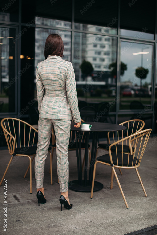 Picture of young lovely caucasian female with dark hair in stylish suit, white shoes stands back to the camera
