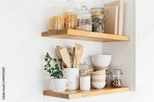 Kitchen shelves with various white ceramic, glass jars, cookbook. Open shelves in the kitchen. Kitchen interior open shelving ideas. Eco friendly kitchen, zero waste home concept