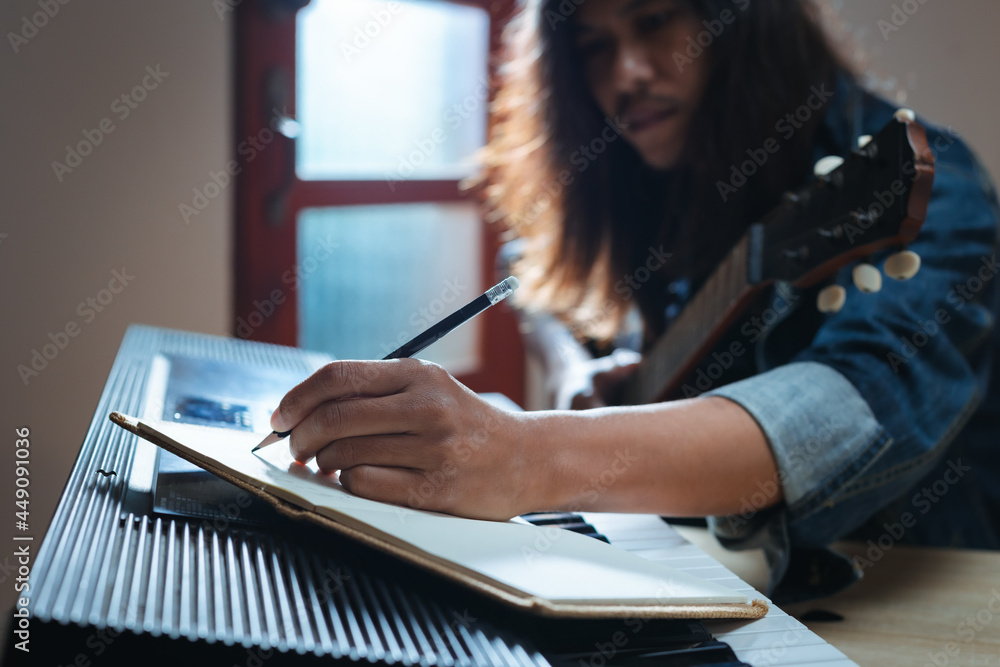 Asian musician who have long hair and wearing jean jacket playing keyboard and guitar and writing a song in notebooks in the dark room and lighting. Asian musician who have long hair and wearing jean jacket playing keyboard and guitar and writing a song in notebooks in the dark room and lighting.