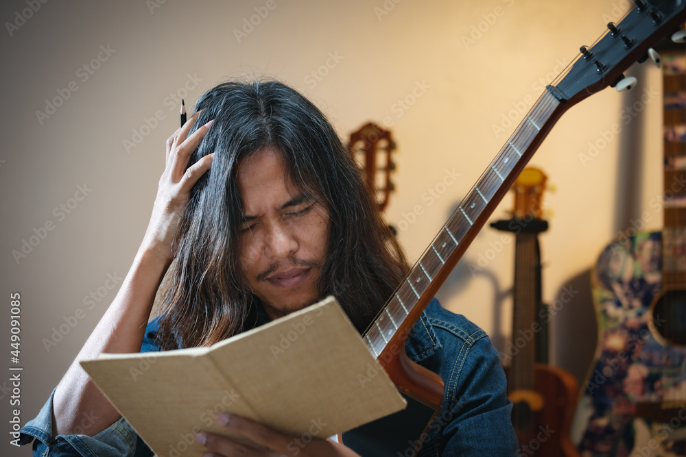 Asian musician who have long hair and wear jean jacket is playing guitar  and so stressed to think about lyric and write a song in notebooks in the  dark room. Photos