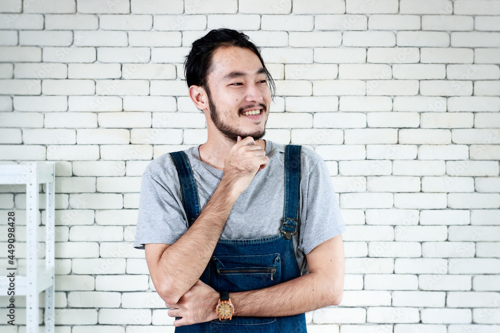 Young man in apron standing and smiling looking to outside in front of white brick wall. Young man in apron standing and smiling looking to outside in front of white brick wall.