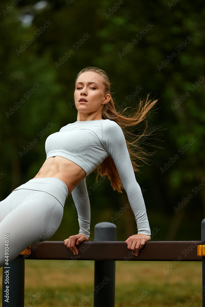 A blonde girl in a tracksuit trains in a park in an urban landscape, a cross-fit instructor doing exercises from yoga, Pilates, deep work, step aerobics.